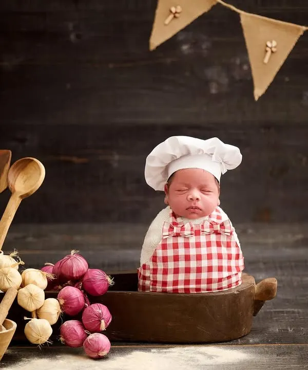 Introducing the world's tiniest chef. This newborn is dressed in a chef's hat and apron, sitting in a cooking pot surrounded by wooden utensils and vegetables.