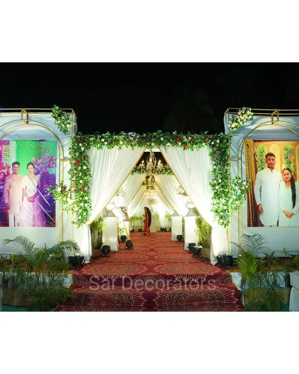 A grand entrance designed to welcome guests in style. This setup features elegant white frames with large couple portraits, a lush green floral arch, and a beautifully draped walkway leading into the venue.