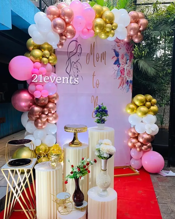 A classic 'Mom to Be' baby shower setup with a pink, rose gold, and white balloon arch. The fluted pedestals and floral accents add a touch of elegance.