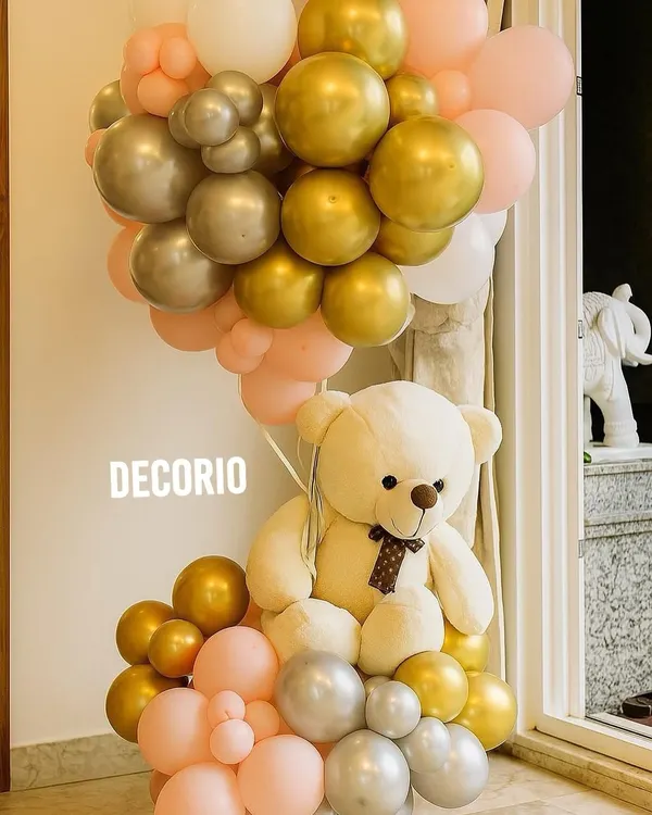 A simple yet beautiful balloon arrangement to welcome a new baby. A large teddy bear is seated within a cluster of pink, gold, and silver balloons, perfect for placing at the entrance of your home.