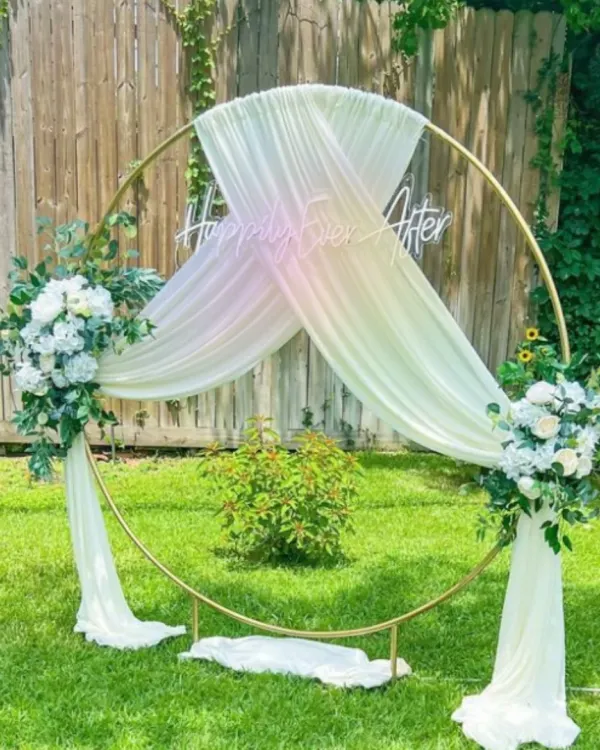 A romantic outdoor backdrop perfect for an engagement or daytime wedding ceremony. The golden ring frame is elegantly draped with sheer fabric and adorned with white floral arrangements, with a "Happily Ever After" neon sign.