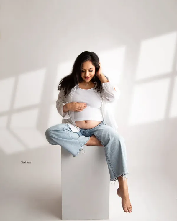 A serene and modern maternity portrait. The mom-to-be is dressed in a simple white top and jeans, sitting peacefully in a brightly lit studio setting.