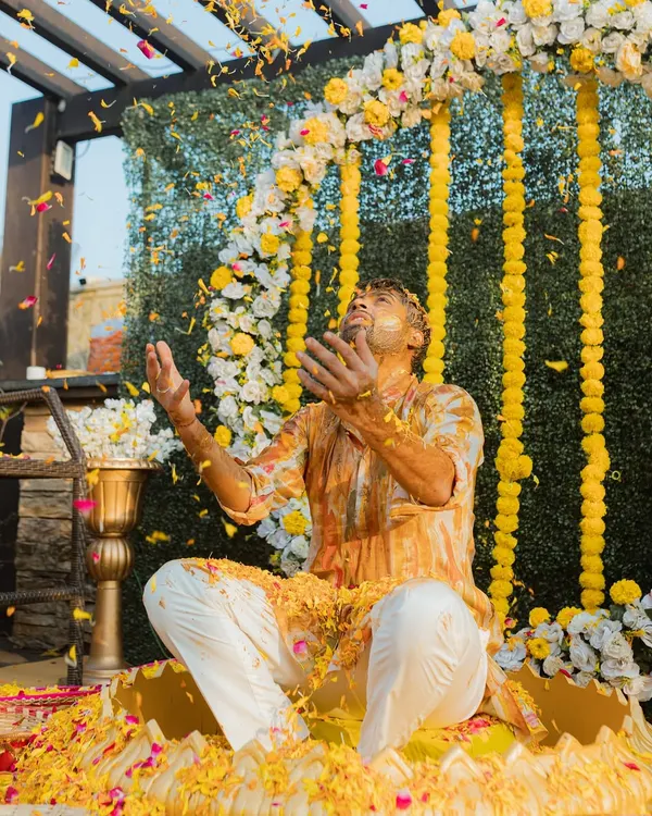 A groom raises his hands in celebration as he is showered with turmeric and flower petals. This action shot perfectly captures the energy of a Haldi ceremony.