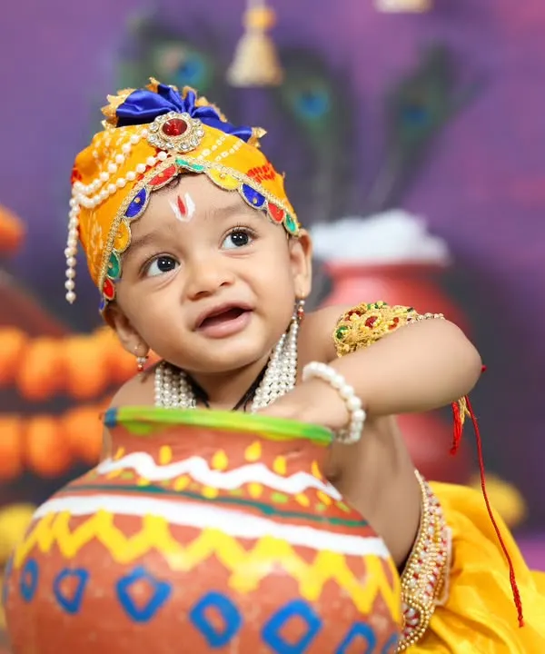 A close-up of a baby dressed as Little Krishna, playfully looking out from behind a butter pot. The detailed costume and traditional props make this a truly divine portrait.