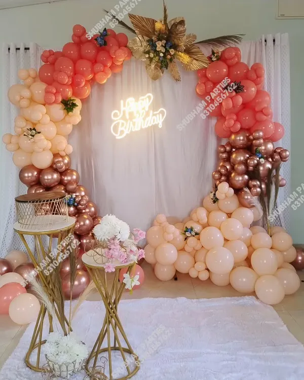 A beautiful boho-chic birthday setup featuring a circular balloon arch with pastel, coral, and rose gold chrome balloons. This elegant party backdrop is completed with a custom "Happy Birthday" neon sign, pampas grass, and stylish cake stands.