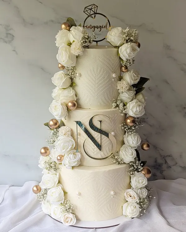 A grand three-tier engagement cake designed for a lifetime of memories. The design features a cascade of fresh white roses and baby's breath, a custom monogram, and delicate stenciling for a look of pure sophistication.