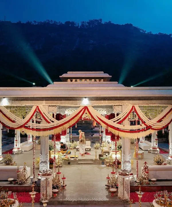 A stunning twilight view of a Kerala-inspired wedding mandap, with the mountains in the background and the structure adorned in traditional red, white, and green florals.