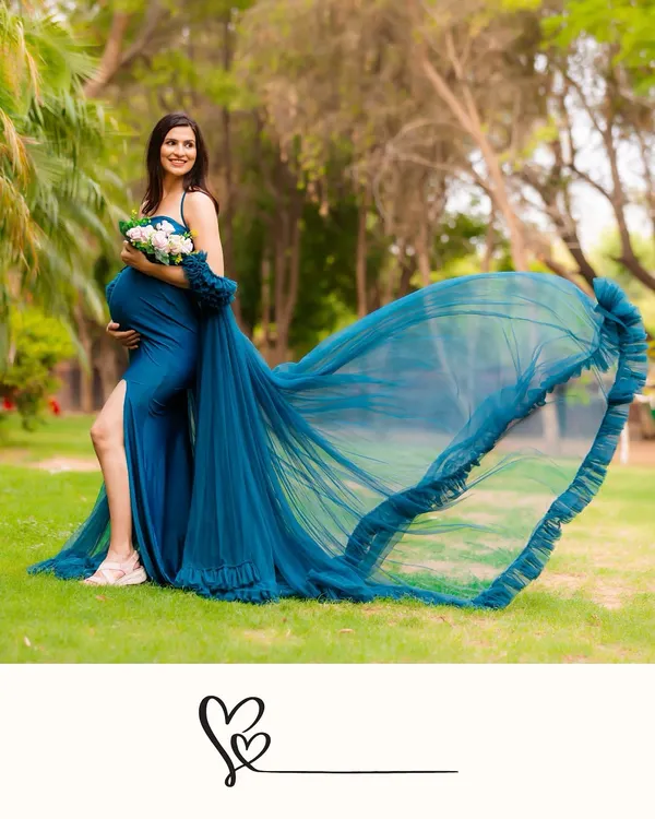 A stunning outdoor shot of a mother-to-be in a vibrant blue gown with a dramatic, flowing train. Holding a bouquet, she smiles, surrounded by the lush greenery of a park.