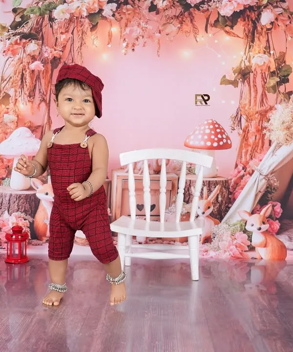 A baby boy stands proudly in a whimsical woodland setup, complete with a teepee, mushrooms, and friendly animal figures. The soft pink and red tones create a warm and magical forest scene.
