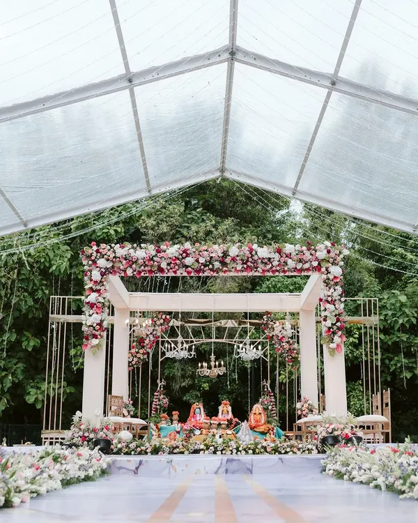 A breathtaking mandap setup inside a clear marquee tent, surrounded by lush greenery. The modern white structure was adorned with beautiful red and white florals and crystal chandeliers for a truly dreamy wedding ceremony.