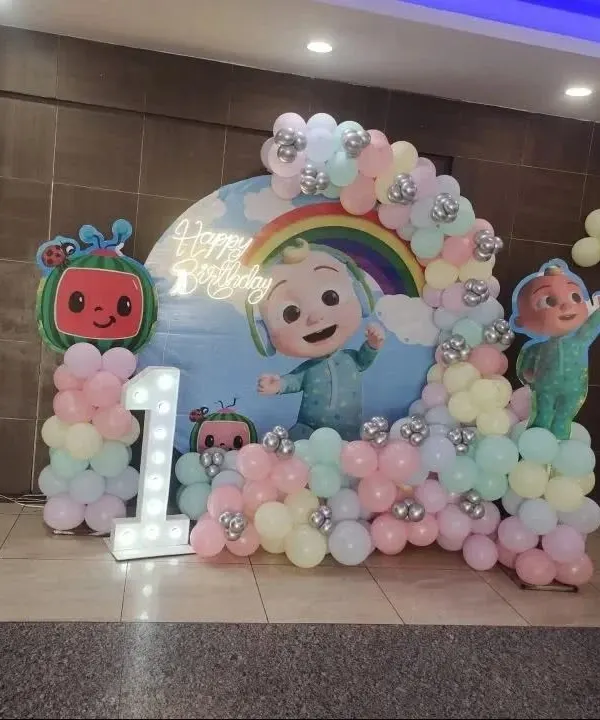 A cheerful Cocomelon theme for a first birthday. This setup features a custom backdrop with a rainbow and Cocomelon characters, a pastel balloon arch, and a large light-up number '1'.