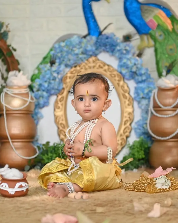 A classic Little Krishna pose. This setup features traditional pots, peacock feathers, and a beautiful arch, creating a perfect backdrop for this baby dressed as Krishna, complete with pearl necklaces.