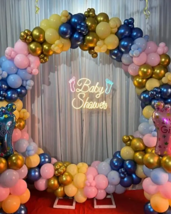 A vibrant gender reveal setup with a circular balloon arch in pink, blue, yellow, and gold. A "Baby Shower" neon sign and "It's a Boy/Girl" foil balloons add to the excitement.