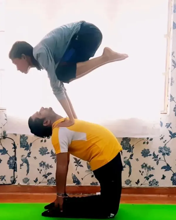 This dynamic acro yoga pose combines the Camel Pose (Ustrasana) and Crane Pose (Bakasana). It requires immense core strength from the top person and a stable base from the partner, showcasing incredible balance and control.