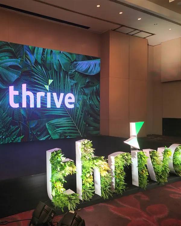 An angled view of the 'thrive' corporate stage. The large LED screen displays branded content, while the custom 3D greenery-filled letters provide a unique, on-theme focal point that is both professional and visually interesting.