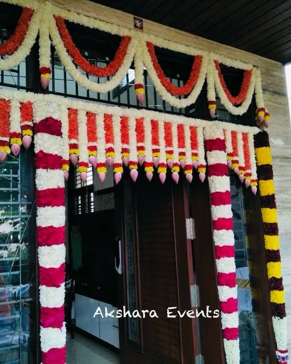A traditional floral decoration for a housewarming ceremony. The main door is beautifully framed with garlands of red, white, and yellow flowers, creating a warm and festive welcome.