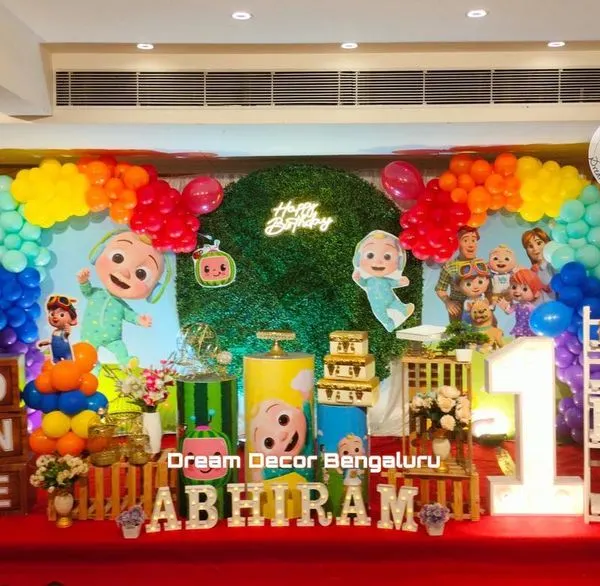 The bright and cheerful world of Cocomelon is perfect for a first birthday party. This setup features a lush green wall, a rainbow balloon garland, and all the beloved characters, including the iconic yellow school bus.