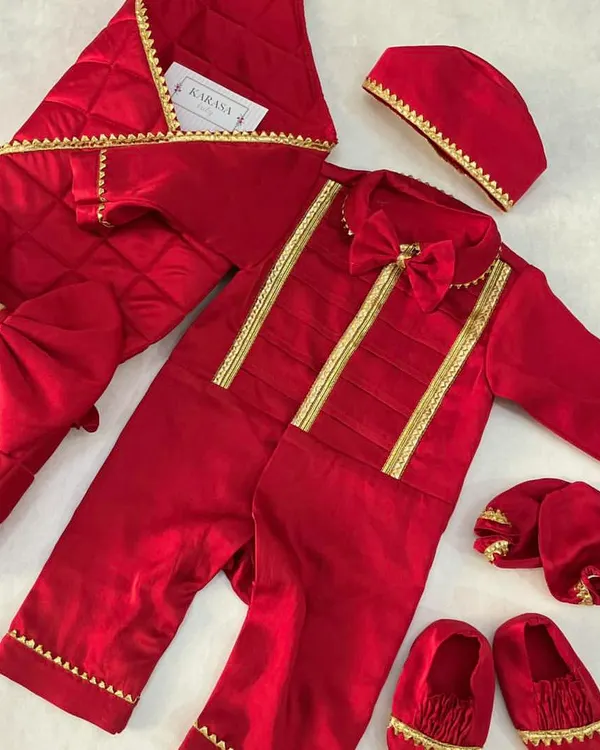A complete newborn Jamna set in royal red velvet with gold trim. This welcoming outfit includes a romper with a bow tie, a quilted blanket, a cap, and matching booties.