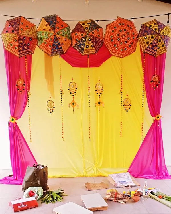 A classic Haldi decor setup with a bright yellow backdrop, pink drapes, and colorful Rajasthani umbrellas. The hanging ornaments add a festive, traditional touch.