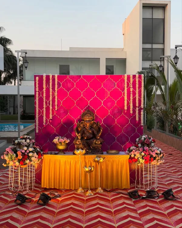 A grand welcome setup for Lord Ganesha, featuring a vibrant pink backdrop with a traditional pattern. The idol is placed on a yellow-draped table, flanked by large floral bouquets and spotlights.