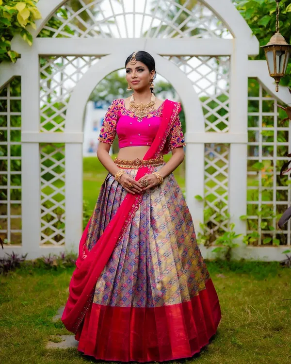 A full-length shot showcasing a vibrant Kanjeevaram half saree. The outdoor garden setting provides a beautiful, natural backdrop that complements the bright colors of the bridal lehenga, ideal for a designer catalog.
