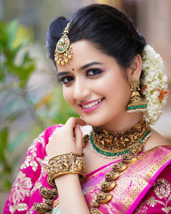 A classic bridal portrait of actress Ashika Ranganath. The focus here is on the detailed temple jewelry and the vibrant pink Kanjeevaram saree, creating a timeless and elegant look.