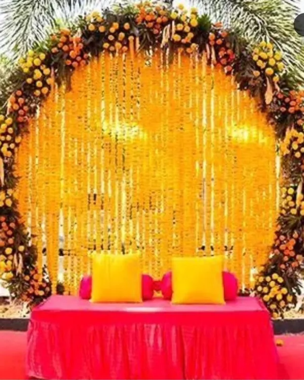 A classic and grand Haldi backdrop featuring a large circular frame completely covered in marigold flower strings. The bright yellow and orange tones create a traditional and cheerful setting for the ceremony.