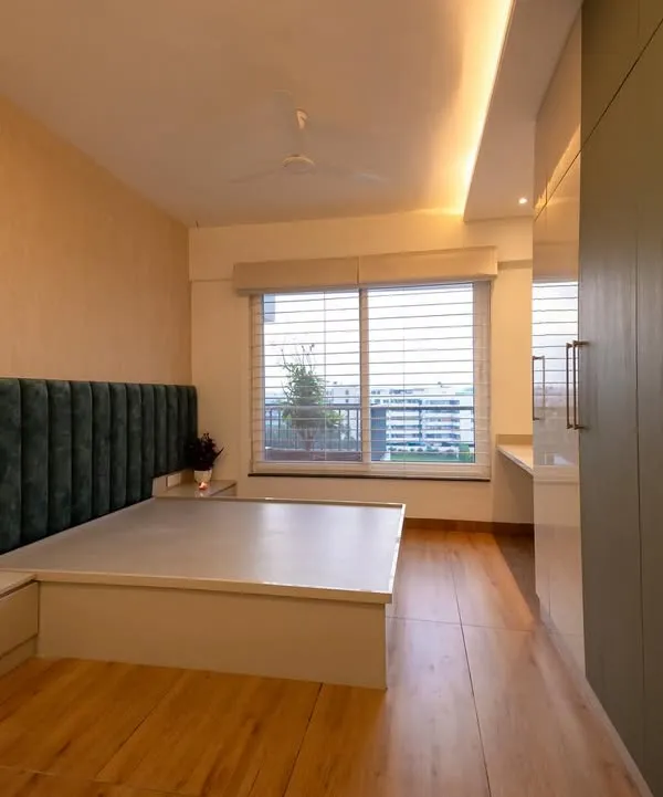 This bedroom is designed for tranquility, with a plush green velvet headboard and a platform bed that includes built-in side tables. The large window with blinds allows for adjustable natural light, creating a calm and restful ambiance.