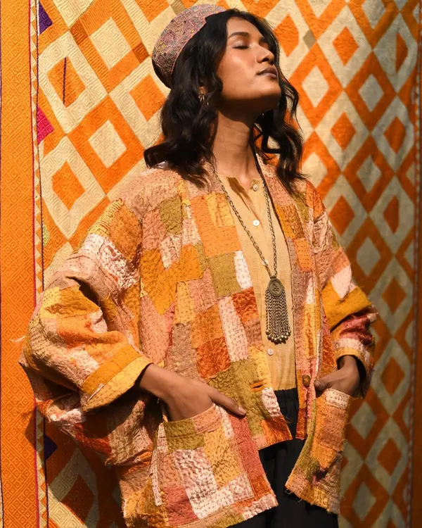 Bathed in warm light against a vibrant, patterned backdrop. This portrait for Wandering Silk captures the rich, earthy tones of a patchwork Kantha jacket, highlighting its texture and artisanal feel.