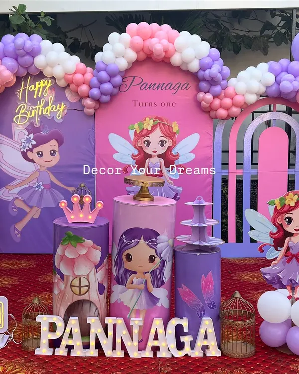 A magical first birthday with a fairy theme, perfect for an outdoor celebration. This setup includes a custom backdrop, a multi-colored organic balloon arch, character cutouts, and themed plinths for the cake and desserts.