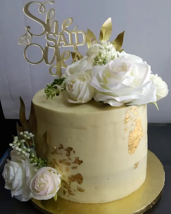 A classic engagement cake for a "She Said Yes" moment. This design features a soft yellow buttercream finish, delicate gold leaf accents, and is topped with beautiful, large white silk roses for an elegant and celebratory look.