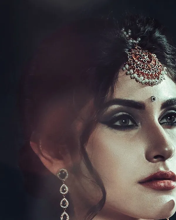 A dramatic close-up where the traditional maang tikka is the hero. The dark, moody lighting is intentional, designed to make the intricate details of the jewelry and the model's smokey eye makeup stand out.