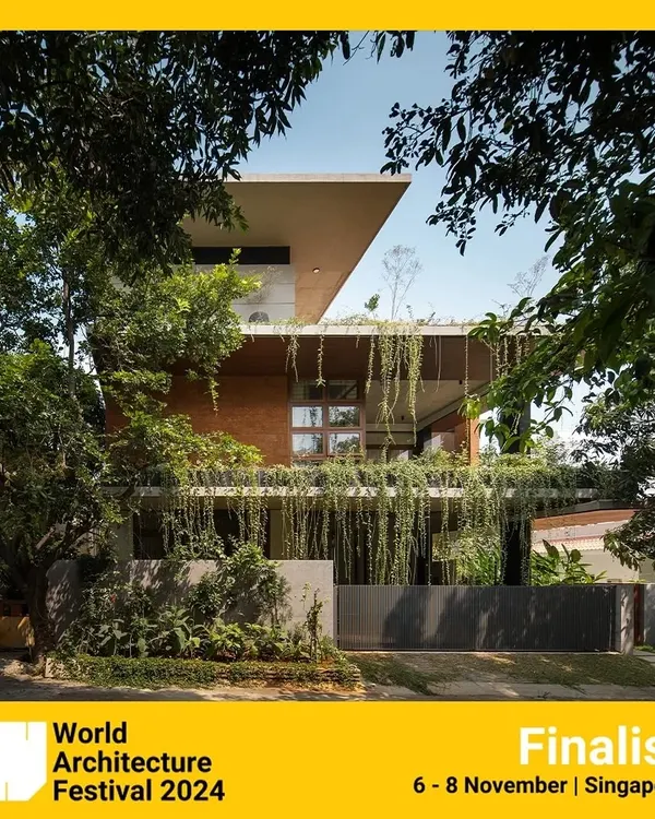 The 'House of Greens' seen from the street, framed by trees. The design uses a combination of exposed brick, concrete, and abundant greenery to create a structure that feels rooted in its natural surroundings.