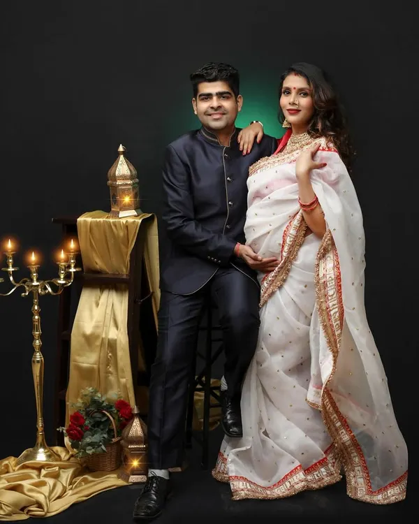 A beautiful couple's portrait celebrating their Indian heritage. The mom-to-be looks stunning in a white and red saree, complemented by traditional props and warm, dramatic lighting.