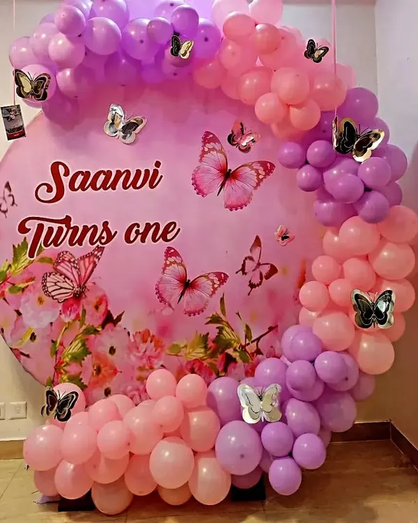 A lovely butterfly theme for a first birthday. The custom circular backdrop reads 'Saanvi Turns One' and is surrounded by a pink and purple balloon arch with decorative butterflies.