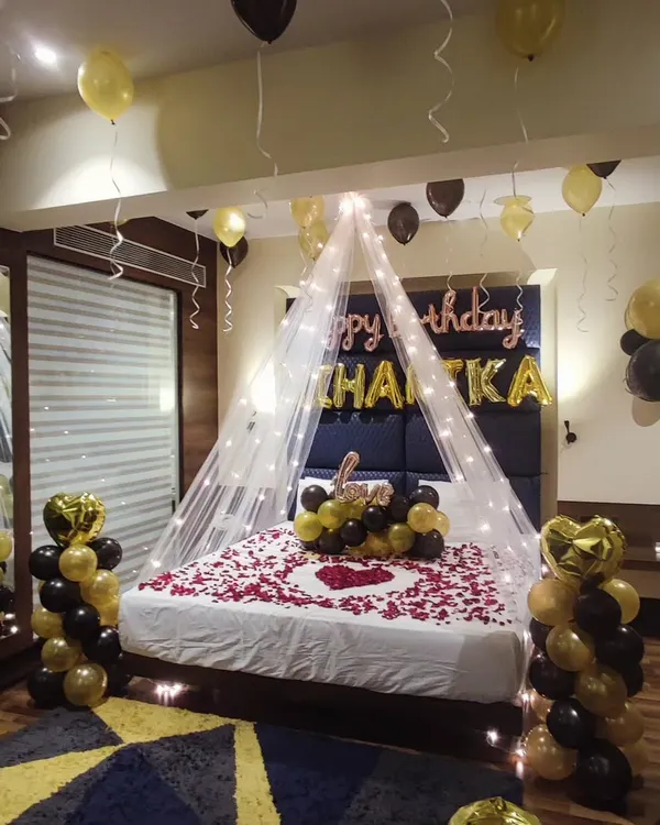 A complete romantic room setup for a birthday. This decoration features a bed canopy with fairy lights, a heart shape made of rose petals on the bed, and black and gold balloon arrangements.