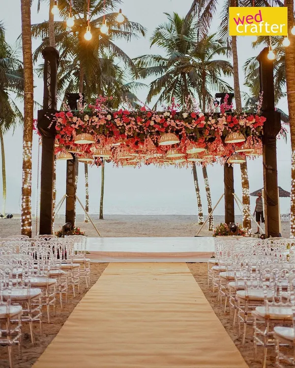 A stunning beach mandap setup facing the ocean. The structure is heavily adorned with pink and red florals, with traditional umbrellas hanging down, creating a perfect frame for a sunset ceremony.