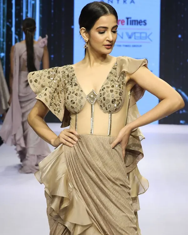This draped saree gown, featured at Bangalore Times Fashion Week, is a showstopper. It has a transparent corset bodice with western-style embroidery and dramatic ruffles on the shoulder and hip, blending tradition with avant-garde design.