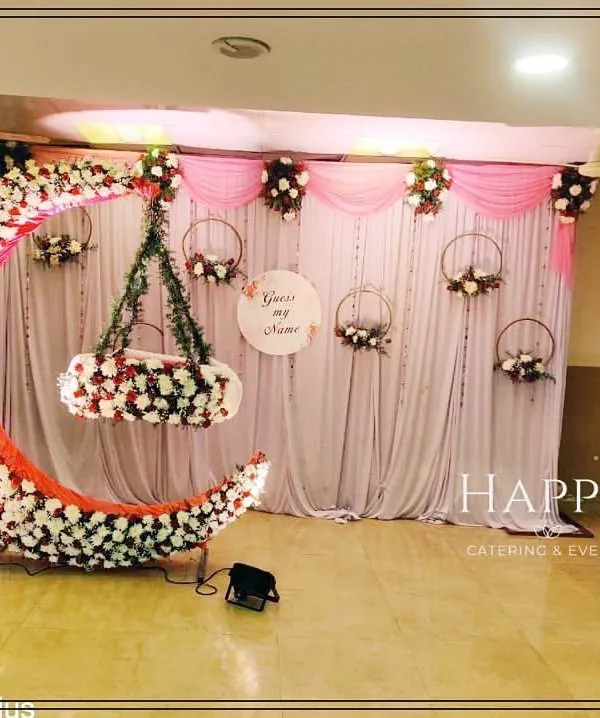 A beautiful crescent moon cradle decorated with white and red flowers, set against a soft pink and white draped backdrop for a naming ceremony. A "Guess my name" sign adds a playful touch.