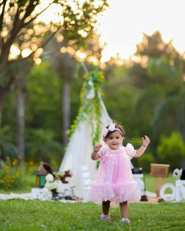 A beautiful outdoor portrait of a one-year-old girl in a pink dress, taking her first steps in a lush green park. The teepee in the background adds a touch of whimsy.