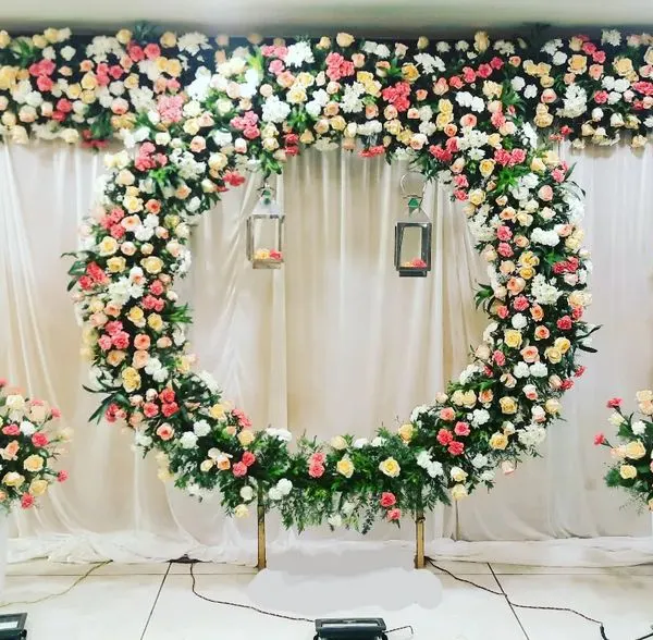This stunning engagement stage features a full ring of multi-colored roses against a clean white backdrop. The symmetrical arrangement of floral bouquets on white pedestals adds to the elegance of the design.