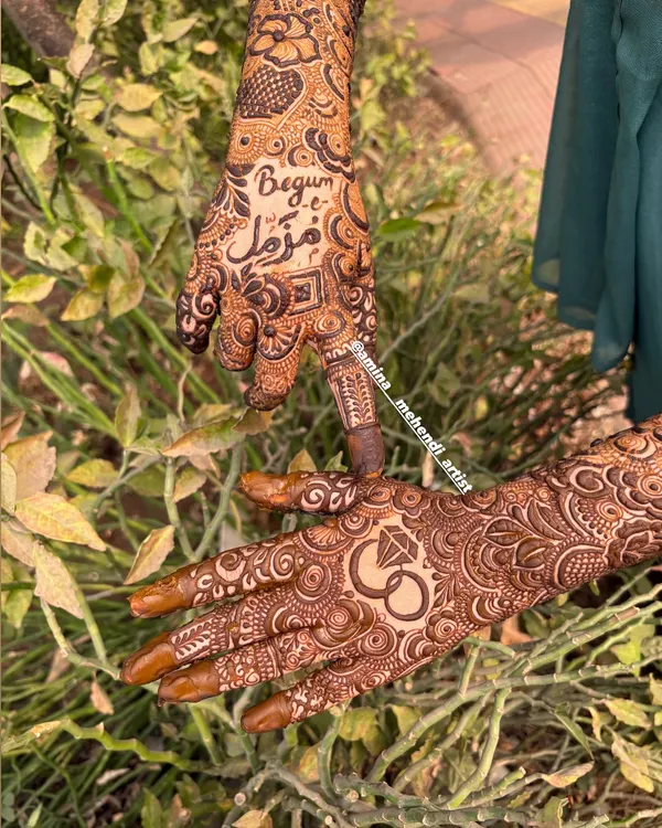 A close-up of a personalized engagement mehendi, featuring the bride's name 'Begum' and the groom's name in Urdu script, alongside a classic engagement ring symbol.