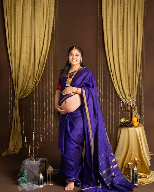A beautiful traditional maternity portrait. The mother-to-be is dressed in a royal blue silk saree, her baby bump peeking out, in a studio setting with classic golden drapes and brass decor.