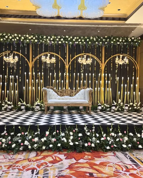 A stunning stage for a Sangeet or Cocktail night. The design features a black and white checkered platform, elegant white seating, and a backdrop of golden arches, candles, and white florals.
