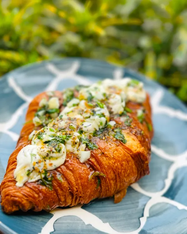 A close-up of our Korean Cream Cheese Croissant. This savory treat is filled with a slightly sweet cream cheese and topped with a delicious garlic herb butter, a must-try from our menu.