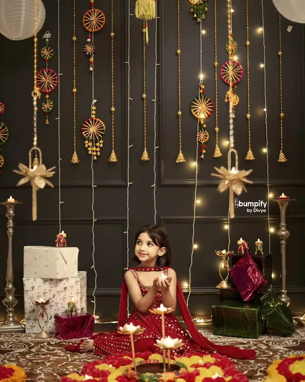 The magic of Diwali through a child's eyes. This little girl in her beautiful red dress is surrounded by the warm glow of diyas and festive decorations in our special Diwali studio setup.