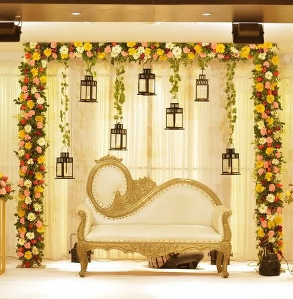 This reception stage design is a perfect blend of modern elegance and classic romance. The floral arch is accented with hanging lanterns that provide a soft glow, beautifully framing the ornate white and gold chaise lounge for the couple.