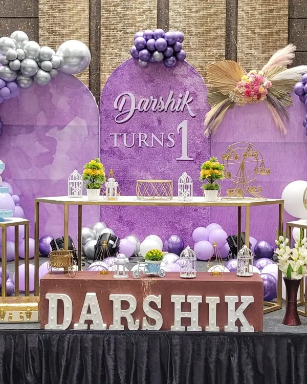 A beautiful first birthday stage in a purple and silver color scheme. The setup includes a custom backdrop, balloon arches, a dessert table, and light-up letters for the child's name.