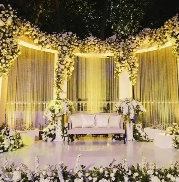 An enchanting outdoor reception stage completely enveloped in white flowers. The cascading floral arrangements and soft, warm lighting create a dreamy and romantic atmosphere under the night sky.