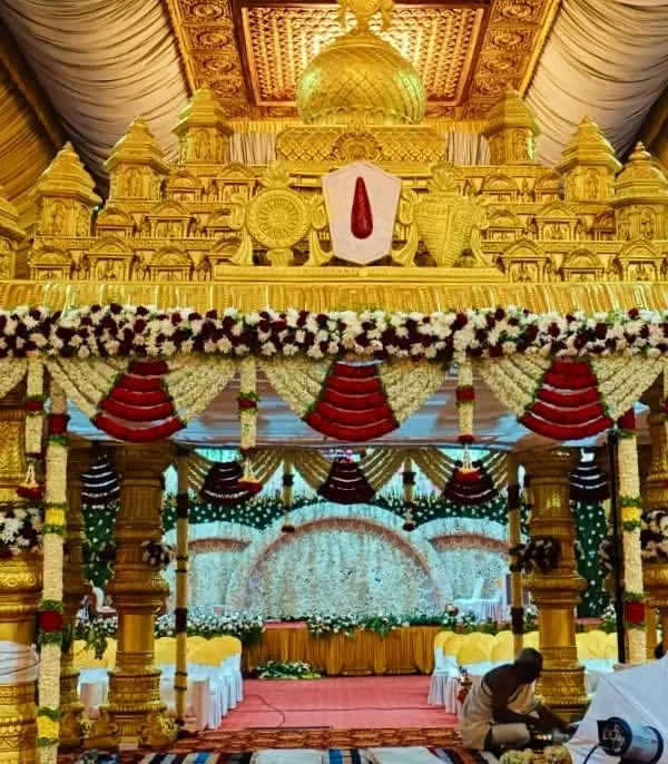 A magnificent golden mandap designed to look like a traditional temple gopuram. This grand setup, decorated with red and white floral garlands, is perfect for a classic South Indian wedding ceremony.
